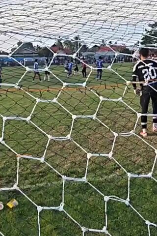 The video shows a soccer game viewed through a soccer net. A goalie in black and white uniform with the number 20 on his back is standing in front of the net. Several other players in blue uniforms are visible on the field. Spectators are gathered in the background. The field appears to be made of grass. There are some discarded items on the grass in the foreground, including a yellow object and some wrappers. The sky is visible above the field, with some clouds. The video appears to be recorded during daylight.