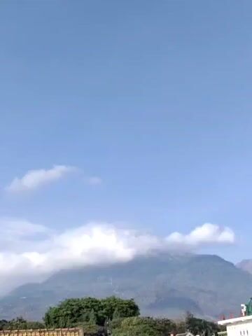 The video shows a scenic view of a mountain shrouded in clouds against a clear blue sky. In the foreground, there are rooftops of buildings and lush green trees. The camera appears to be stationary, capturing a still image of the landscape. The lighting suggests it is daytime, possibly morning or afternoon. There are no people or active events depicted in the video.
