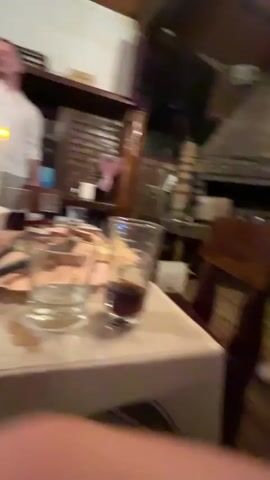 The video shows the interior of a restaurant. A woman in a short, white strapless dress stands near a brick fireplace, talking to another woman who is partially visible. There is a table with a glass of liquid in the foreground. There's also a wooden chair and what looks like a kitchen area with various objects. The scene is dimly lit with a warm, rustic ambiance.