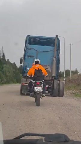 A person on a motorcycle, wearing a white helmet and an orange vest, rides behind a large blue semi-truck on a dirt road. The motorcycle's license plate reads "8419 AH 59". The truck has a large exhaust pipe on its right side. The road is lined with trees and vegetation on both sides. The sky is overcast.