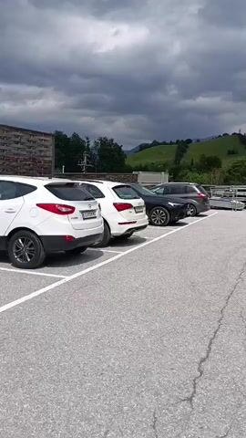 The video shows a parking lot with several cars. The cars are parked in designated spots. In the background, there are green hills and a cloudy sky. There are no people visible in the video. The overall scene appears to be calm and quiet.
