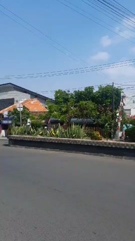 The video shows a street scene with several buildings, a statue, and power lines. The buildings appear to be commercial establishments, with signs and storefronts visible. One building has "QUINA" written on it. There is a statue of a person in traditional clothing, with arms outstretched. Several people are seen on motorcycles, and there are trees and plants along the side of the road. The sky is clear and blue, with some clouds. The video appears to be filmed from a moving vehicle, as the perspective shifts and the scenery blurs slightly.