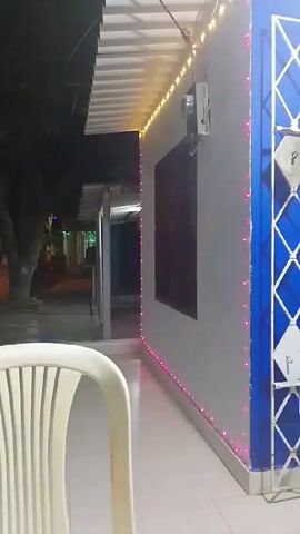 The video shows a night scene of a street in what seems to be a residential area or small town. A white plastic chair is in the foreground, partially obscuring the view.  The camera pans to show the street with houses decorated with festive lights. The decorations are multicolored LED strings outlining windows, doors, and roofs. Trees are present on the street, adding to the nighttime ambiance. In the lower part of the shot, there's a red container with some objects inside, which could be craft supplies. The scene is peaceful, lit by the festive lights and some streetlights. The music in the background adds to the festive atmosphere.