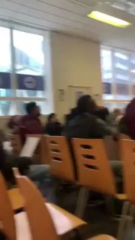 The video shows a room full of people sitting in chairs, facing forward. The chairs are light brown and have cut-out designs. The room is lit by fluorescent lights. There are large windows in the background, revealing a view of buildings outside. The people are of various ages and ethnicities. Some are wearing head coverings. The video appears to be taken from a low angle, moving slightly.