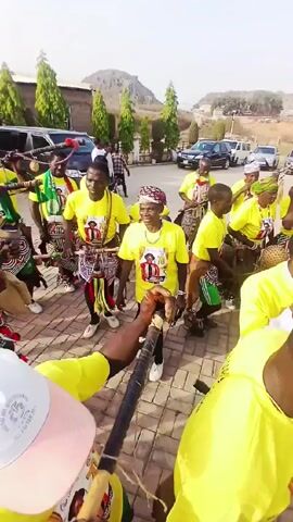 A group of people in yellow t-shirts are dancing and playing musical instruments on a bricked surface. The instruments appear to be horns, drums and stringed instruments. Several cars are parked in the background. There is a modern looking building and trees in the background.  The people are smiling, and appear to be enjoying themselves.