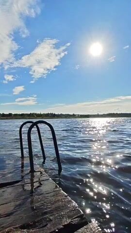 The video shows a close-up view of a wooden dock on a body of water. A metal ladder with two arched handles is partially submerged in the water. The water is choppy and reflects the bright sunlight. The sky is blue with scattered white clouds. In the distance, a treeline is visible. The sun is high in the sky and appears as a bright, white orb. The scene is calm and serene, evoking a sense of a warm summer day.