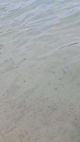 The video shows a calm, clear body of water, likely a beach or cove. In the foreground, the water is shallow enough to see the sandy bottom. Moving towards the horizon, there are people swimming, some closer to the shore and others further out. Several boats are anchored off the coast. The background features a hilly coastline covered in vegetation. The sky is partly cloudy with patches of blue visible. The overall scene is a tranquil beach setting with people enjoying the water.