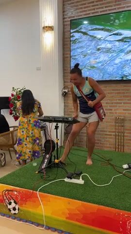 A woman with dark hair and a yellow floral dress stands to the left, facing a keyboard. To the right, a woman with her hair in a bun, wearing a blue and teal tank top and gray shorts, plays a drum pad with drumsticks. She is barefoot and has a red and white bag slung across her body. Both women are on a raised platform covered in artificial grass. The background features a brick wall, a large television displaying moving water, and a decorative floor lamp. Wires and power strips are visible on the platform. The overall setting appears to be an indoor event or performance space.