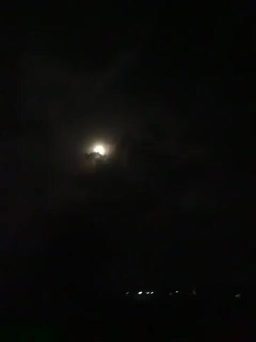 The video shows a nighttime scene with a dimly lit moon partially obscured by clouds. In the distance, faint lights suggest a populated area. The camera appears to be moving or is handheld, causing slight instability.
