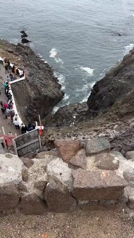 The video shows a high-angle view of a crowd of people gathered on a cliff overlooking the ocean. The cliff is rocky, and there are waves crashing against the rocks below. There is a blowhole that appears to be ejecting water. There are buildings and tents on the cliff near the crowd. The sky is overcast.