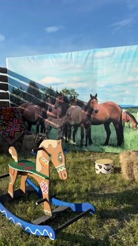 A wooden rocking horse with blue rockers sits on a grassy area. Behind the rocking horse is a backdrop of horses in a field. To the left of the rocking horse is a wooden fence and some hay bales.