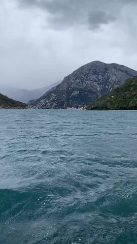 The scene shows a body of water, likely a bay or lake, with mountains visible in the background. The water is a deep blue-green and has some waves, indicating a bit of wind. The mountains are grey and rugged, with some patches of green vegetation. The sky is overcast and grey, suggesting a cloudy or rainy day. In the distance, there appears to be a small village or town nestled along the shoreline. The view is taken from a low angle, possibly from a boat on the water.