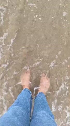 The video shows a pair of bare feet standing on a sandy beach with the ocean water gently lapping around them. The camera is positioned directly above the feet, looking down. The person is wearing blue jeans that come down to their ankles. Small shells and pebbles are scattered on the sand. The water is clear and shallow, with small waves creating ripples around the feet and leaving foamy trails on the sand. The scene is calm and serene, suggesting a relaxing moment by the sea.