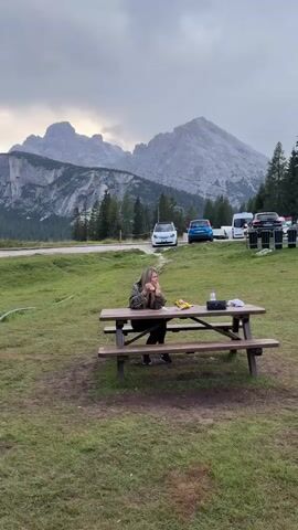 A young woman sits at a picnic table in a grassy area with mountains in the background. She is wearing a leopard print jacket and black pants. There are bags of snacks and a water bottle on the table. She is looking up and to the side, with her hands resting on her chin. Cars are parked in the background. The sky is cloudy.