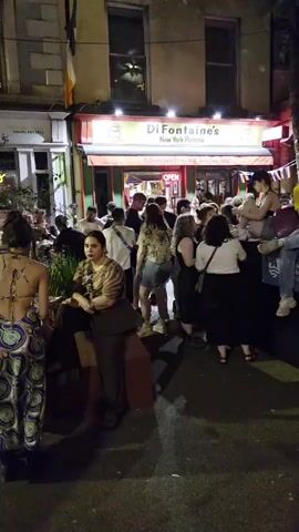 A crowd of people are gathered outside a pizza restaurant called Di Fontaine's New York Pizza at night. A large, brightly colored statue resembling a fox, painted with rainbow colors and other symbols, stands to the right of the entrance. Some people are sitting on a low wall in front of the statue. Several individuals are looking at their phones. Others are standing and talking. The restaurant has its door open and lights on. There are flags and banners displayed on the building. The street is paved and appears to be a public area. The overall atmosphere seems to be lively and social.