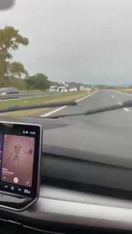 A video depicts a car driving on a highway on a cloudy day. The view is from the driver's perspective, focusing on the road ahead and the car's interior. Cars and trucks can be seen on the highway in the distance. The car's dashboard is visible, displaying a navigation system or entertainment unit playing music with the cover art of a song called "Bones" by Imagine Dragons. Road signs, including directional and no-smoking signs, are visible along the road. The car appears to be driving on a wet road surface, with occasional raindrops on the windshield.