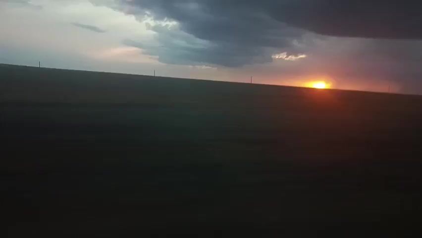 The video shows a wide shot of a vast, dark field under a dramatic sky at dusk. The horizon is dimly lit by a setting sun, which appears as a bright orange ball peeking over the distant, flat landscape. Dark, heavy clouds dominate the upper portion of the sky, with a few lighter clouds scattered near the sun, tinged with pink and yellow hues. The field itself is almost entirely obscured by shadow, appearing as a dark, blurry expanse. A few faint, vertical lines, possibly power poles or wind turbines, are sparsely distributed along the distant horizon. The overall impression is one of a desolate, rural landscape bathed in the fading light of day. The camera appears to be stationary or moving very slowly.