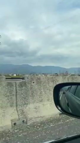 The video is taken from inside a car, likely in the passenger seat, looking out the side window. The car is moving along a highway, with a concrete barrier separating the highway from a town beyond. The scenery includes buildings, trees, and distant mountains under a cloudy sky. In the car's side mirror, the back of another car is visible.