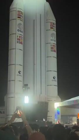 The video shows a crowd of people in front of a large model of a rocket at night. The rocket is white with the ESA and CNES logos on it. The crowd is facing a stage set up in front of the rocket. People in the crowd are holding up their phones, presumably recording the event. Lights are shining on the stage and the crowd. The environment is dark. The video is shot from the crowd's perspective.