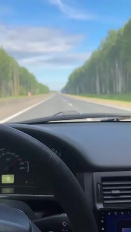 The video provides a first-person perspective from inside a car, looking forward through the windshield while driving on a highway.

**Subjects and Objects:** The main subjects are the car's interior elements and the road ahead. In the foreground, a black steering wheel is prominent. Behind it, the dashboard is visible, featuring a speedometer on the left with the needle fluctuating between approximately 80 and 90 km/h. Below the speedometer, the odometer displays "190926" (kilometers), and a trip meter shows "163.9." Several indicator lights are illuminated on the dashboard, including a yellow warning light. To the right, an aftermarket infotainment screen shows a blue and white "Bluetooth" connection interface. A rearview mirror hangs from the ceiling.

**Scene and Setting:** Through the windshield, a straight, multi-lane asphalt highway stretches into the distance. The road appears well-maintained, with clear white lane markings and a concrete barrier or grass median separating the directions of travel. Both sides of the highway are lined with dense, tall forests, primarily composed of birch and other deciduous trees, creating a green "tunnel" effect. The sky above is bright blue with scattered white clouds.

**Activity/Event:** The car is actively being driven down the highway at a steady speed.

**Notable Details:**
*   **Time of Day:** It is clearly daytime, with bright sunshine.
*   **Weather:** The weather is clear and sunny.
*   **Location Cues:** The presence of extensive birch forests is common in regions like Russia. The speedometer readings are in kilometers per hour.
*   **Sounds:** Music is playing in the background throughout the video.

The overall impression is one of a calm, continuous journey on a rural or intercity highway.
