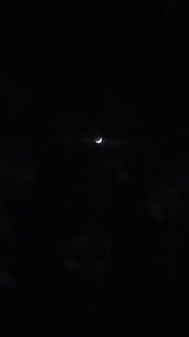 A very dark video shows a crescent moon in the center of the frame. The moon is faintly illuminated and surrounded by a dark sky with some cloudy or hazy areas. The video is grainy and appears to have been taken at night.