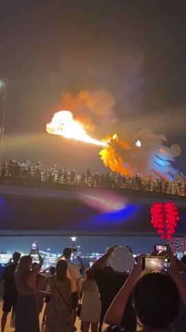 At night, a white and blue dragon sculpture is depicted blowing fire from its mouth. The dragon is positioned on a bridge above onlookers, some of whom are taking photos. The area below the bridge is illuminated with blue light. Red lantern decorations hang below the bridge on the right side of the video. The sky is filled with smoke from the fire.