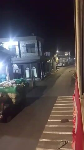 The video captures a nighttime view from a moving vehicle, likely a train, looking out a window. The scene outside is a street with buildings on the left and right, illuminated by streetlights and lights from the buildings. On the left side of the street, there are large piles of goods wrapped in green and white plastic sheeting, stacked on what appears to be a vehicle. A two-story building with a patterned facade is visible behind these goods. The street itself is paved and slopes slightly uphill. On the right side, a person's foot wearing a sandal is visible on a set of stairs or a platform. The train appears to be moving slowly. The overall atmosphere is dark and residential.