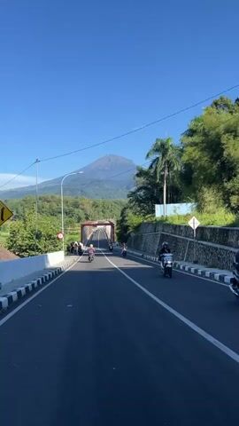 The video shows a road leading to a red bridge, with a large mountain in the background under a clear blue sky. The road is paved and has white lines down the center. There are black and white striped curbs on either side of the road. Several people are standing on the left side of the road near the bridge. Several motorcycles are driving towards and over the bridge. The environment is green with trees and other vegetation. There is a stone wall on the right side of the road. A sign is visible on the left side of the road.