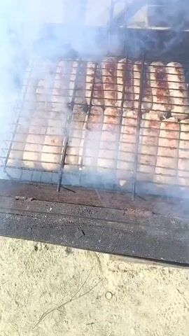 The video shows sausages grilling on a barbecue. The sausages are arranged on a wire rack over the grill. There is smoke rising from the barbecue. In the background, there is a table, a cauldron on a stand, a log cabin-style house with a covered porch, and a black car parked in the driveway. The setting appears to be a backyard or outdoor area, possibly near a residential home. The weather is sunny and clear.