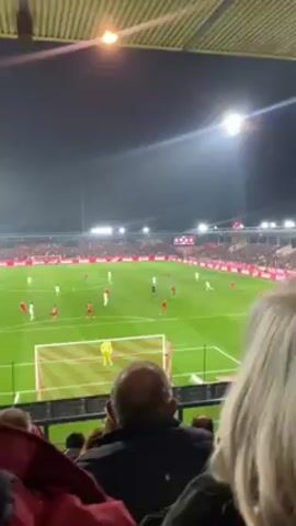 The video shows a view from the stands at a soccer game. The perspective is from behind spectators, partially obstructing the view of the field. The stadium is packed, with fans filling the seats. Players in red and white uniforms are on the green field. The lights are on, illuminating the game. The camera angle is relatively steady, providing a clear view of the field and surrounding stadium.