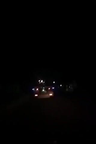 A vehicle is driving on a dark road at night. The camera is behind the vehicle, capturing the taillights and occasional light from streetlights or other sources. The road appears empty.
