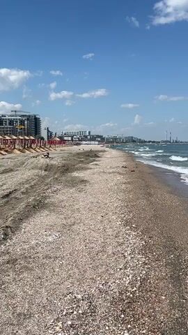 The video shows a beach on a sunny day. The foreground is a gravelly beach with a few pieces of trash. The sea is blue with white waves. In the background, there are buildings and industrial structures. A few people are visible in the water. The sky is blue with some white clouds.