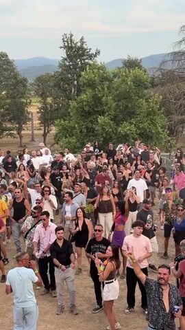 The video opens with a wide shot of a large crowd of people standing in an outdoor setting, some with their hands raised. The crowd is densely packed, with individuals of various ages and genders. The environment appears to be a festival or outdoor event, with trees and hills visible in the background. The scene transitions to a close-up shot of a DJ on a stage. The DJ, a male with dark hair, is wearing a black t-shirt and jeans and is actively engaged in mixing music, with his arm raised. He is positioned behind a DJ booth with speakers and lighting equipment surrounding him. The overall impression is of a lively and energetic outdoor music event.