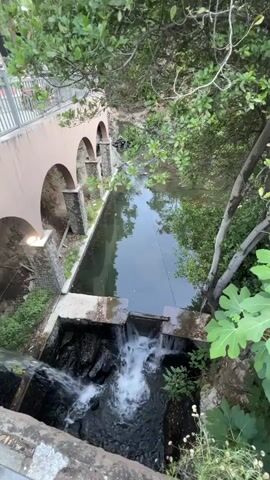 This is a video of a small waterfall and stream in a setting with lush greenery. A light pink arched structure is visible to the left, with stone pillars supporting arches. The stream flows into a small plunge pool where it cascades over rocks. The water is clear and reflects the surrounding trees and sky. Sunlight filters through the leaves, creating dappled patterns on the water and ground. The overall impression is one of tranquility and natural beauty. The video appears to be taken during the daytime. No people are visible in the video, nor is any text. The location context provided is Kakopetria, Cyprus.
