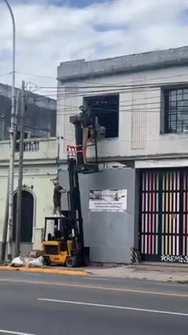 The video shows two men lifting an object into a second-story window with a forklift. A man is standing on the forklift behind the driver controls, apparently guiding the lift. Two men are visible in the window, reaching to receive the object being lifted. The forklift is yellow and black. The building is white with a grey panel over which is taped a sign. The doors to the right are painted with vertical stripes. There is a utility pole with electrical wires to the left of the building. A street runs in front of the building.