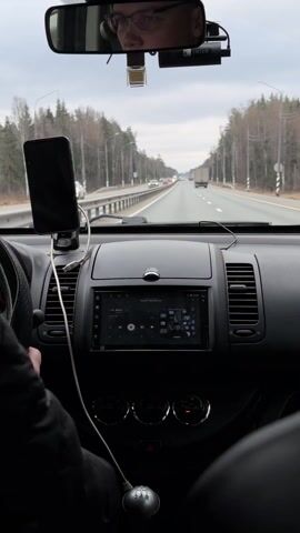 A person is driving a car on a highway. The driver wears glasses and is reflected in the rearview mirror. A smartphone is mounted on the dashboard, connected to the car's multimedia system with a USB cable. The system's screen displays a media player interface. The car appears to be a Nissan Note with a manual transmission. Outside, the sky is overcast, and trees line the sides of the road. Other vehicles are visible in the distance.