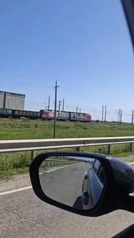 The video shows the view from a car moving on a road, with a train running parallel to the road. The train is red and gray and appears to be carrying cargo. The road is asphalt and has a guardrail. The sky is blue and clear. The car's side mirror is visible in the lower left corner of the frame. There is green grass and other vegetation between the road and the train tracks.  Electrical wires and poles run along the train tracks.