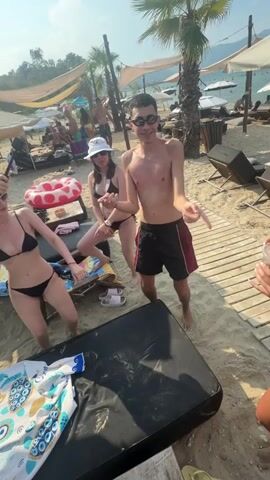 A group of young adults are dancing on a sandy beach. The person in the center foreground, wearing black shorts and no shirt, is performing a dance with hand gestures. To their left, a woman in a black bikini is also dancing, holding a phone in her right hand. To the center left, another woman in a black bikini and white bucket hat is dancing. In the background, other beachgoers are visible, with umbrellas and lounge chairs set up on the sand. The overall atmosphere appears to be festive and summery.