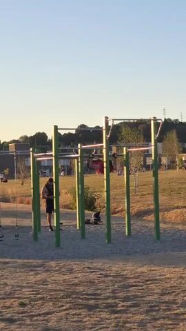 A person is upside down on a horizontal bar. They are wearing a red shirt and dark shorts. Their hands are gripping the bar. In the background, there is a park with trees and grass. Another person is standing to the left of the bar, looking down at their phone.