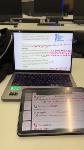 The scene shows a laptop and a tablet on a desk in a room with multiple computer monitors in the background. The laptop screen displays a document with notes written in red pen. The tablet screen shows a form with checkboxes and fields filled with handwritten notes. The environment appears to be a classroom or office setting with multiple workstations. No people are visible in the scene. The focus is on the electronic devices and their screens.