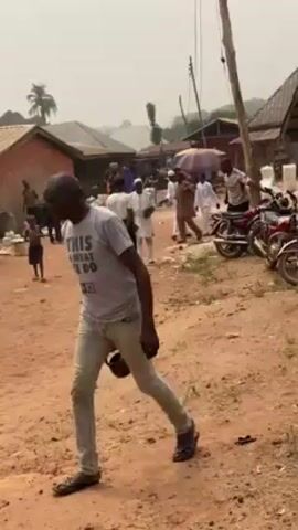 The video shows a dirt road in what appears to be a small village or town. There are several people walking around, some wearing traditional Muslim clothing, and others in more casual attire. Some individuals seem to be conducting business or selling goods by the roadside, with an umbrella providing shade over some wares. Several motorcycles are parked nearby. The environment looks somewhat dusty or hazy, and the buildings are modest and simple in construction. Overall, the scene depicts everyday life in a rural or less developed area.