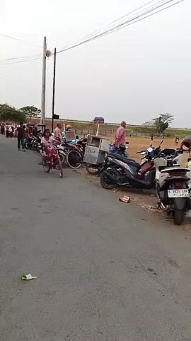 A young girl rides a pink bicycle on a paved road. She is wearing a pink shirt and patterned pants. Behind her, several motorcycles are parked along the side of the road. In the background, a grassy field can be seen, where a group of people, some wearing yellow shirts, are gathered. There are also trees and a utility pole visible in the distance. The sky is overcast.