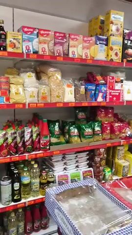 The video shows the interior of a small grocery store. Shelves are stocked with various food items, including packaged goods, snacks, and beverages. A refrigerated display case in the foreground contains cheeses, dairy products, and some packaged meats. A person wearing a grey hoodie and blue gloves is visible, reaching into the refrigerated case. The store appears to be well-lit and organized. The overall impression is that of a typical neighborhood convenience store.