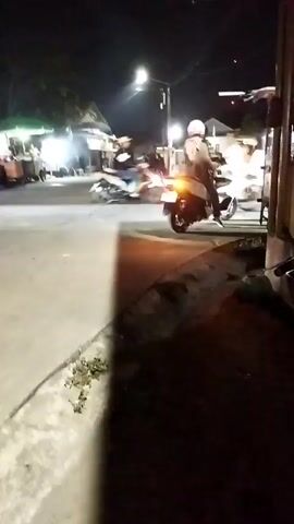 The video captures a nighttime street scene, likely in a commercial area with several stalls or small businesses visible. The lighting suggests it's after sunset, with artificial lights from streetlamps and the stalls illuminating the area. In the foreground, a concrete sidewalk with some sparse vegetation is visible. The main action unfolds on the paved street where multiple motorcycles are present.

One motorcycle, a white scooter-type, is parked on the right side of the frame, facing left. The rider, wearing a helmet and a light-colored jacket, is on the motorcycle. Further down the street, another motorcycle, also a scooter type, is in motion, its taillight illuminated, indicating movement. This motorcycle has at least two occupants, one of whom is wearing a helmet.

In the background, several stalls are set up. One stall on the left has a green awning and appears to be serving customers. People are gathered around this stall, some standing and some seated. Another stall to its right has a blue awning and also seems to have people present. Further back, more structures and lights suggest a more developed area beyond the immediate scene. The overall atmosphere appears to be lively, with activity suggesting a marketplace or a gathering spot. The movement of the motorcycles and the presence of people at the stalls indicate a busy evening.