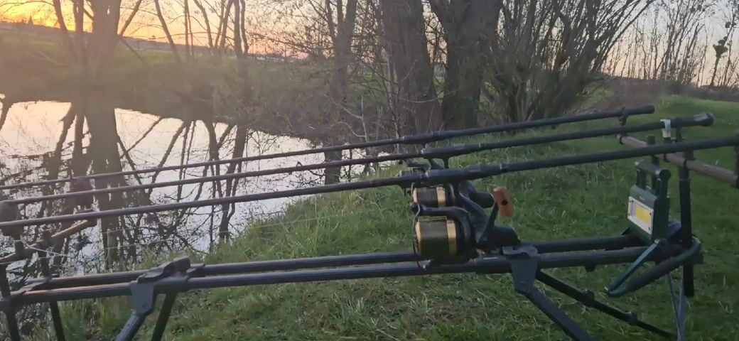 The camera pans up and to the left showing a tranquil scene. In the foreground are three fishing rods set up on a black rod stand. The rods are black and the reels appear to be gold. The rods are positioned near the edge of a body of water, possibly a lake or a wide river. The water's surface is calm and reflects the surrounding trees and the colors of the sky. The sky is transitioning with a soft orange glow on the horizon, indicating either sunrise or sunset. The banks of the water are lined with trees, their bare branches creating a stark silhouette against the colorful sky. There is green grass visible. In the distance, a few hills are visible.