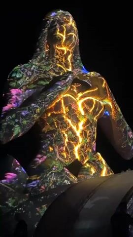 A video shows a projection mapping performance on a statue. In the first shot, the statue is lit with an orange, yellow, and green projection that resembles lava and floral patterns. The statue appears to be a female figure in a seated position, with her hands forming a heart shape over her chest. In the subsequent shots, the projection changes to a more abstract light show with soft pinks and blues, and then shifts to a solid red light that obscures the details of the statue. The background is dark, suggesting a theatrical or performance setting.