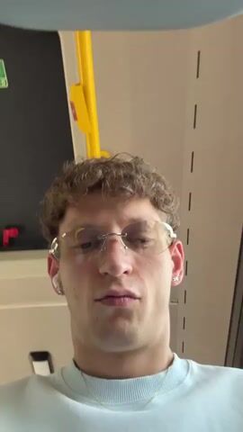 The video begins with a close-up, slightly blurry shot of a young man wearing glasses and a light blue t-shirt. He appears to be inside a vehicle, possibly a bus, as indicated by a yellow pole and a green emergency exit sign visible in the background. The man's head is tilted slightly, and he is looking forward. The camera then pans to the right, capturing a fast-moving view of the scenery outside the vehicle. Trees and a highway are visible, suggesting the vehicle is in motion. The shot is blurry due to the speed and motion. The video ends with another quick shot of the scenery, showing more trees and a grey car driving on the highway.