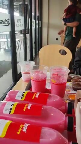 The video begins with a close-up of several bottles of pink liquid in plastic bottles, with red labels that say "Taste Factory." In the foreground, there are also cups of pink drinks with ice, and small containers of sauces and condiments. In the background, a person is holding a child. The camera then pans to the left, revealing more of the table with trays of fried chicken in red and white boxes, as well as more sauce containers and orange jelly cups. To the right of the fried chicken, a woman wearing a hijab and glasses is looking at her phone. The camera continues to pan, showing more tables and chairs in a restaurant setting. There are other people dining in the background, and the restaurant appears to have wooden tables and chairs, with a light-colored ceiling and wooden beams. A poster with a hamburger image is visible on the wall. The video concludes with a wider shot of the dining area, showing several tables occupied by customers.