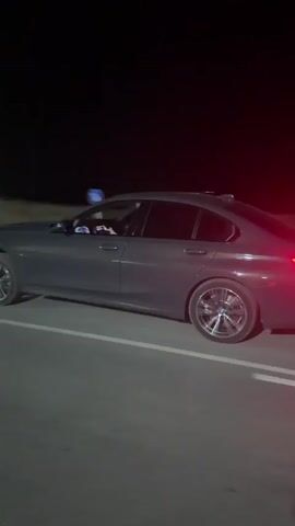 A dark gray sedan, possibly a BMW 3-series, is captured in motion on a two-lane road at night. The vehicle is moving away from the camera, its taillights illuminated and casting a red glow on the asphalt. The scene appears to be on the outskirts of a town or rural area, indicated by the presence of streetlights, utility poles, and sparse vegetation along the roadside. Traffic signs are visible, including a round sign with a red border and a horizontal bar, suggesting a prohibitory measure, and a blue circular sign with a bicycle icon, indicating a bike lane or route. The road is marked with white lines, and there are double white lines in the center, indicating the direction of travel. The sky is dark, suggesting it is nighttime, and there are no indications of weather conditions other than the clear darkness. The overall atmosphere is one of a quiet, possibly late-night drive. The specific location context of Koprinka, Bulgaria, is provided, but not visually evident within the frame itself beyond the general appearance of a European road. No people are visible in the video. There is no discernible text on the car, though a license plate is visible. There is also some text on the traffic signs, but it is not legible.