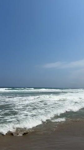 The video shows a beach scene with the ocean and a clear blue sky. The camera is positioned at a low angle, looking out towards the water. Waves are breaking on the shore, creating white foam. The water is a mix of blue and green, and the sky is a vibrant blue with a few wispy clouds. There are no people or objects clearly visible in the foreground, but a few distant figures appear to be in the water. The sand on the beach is a light brown color. The overall impression is one of a peaceful and serene day at the beach.